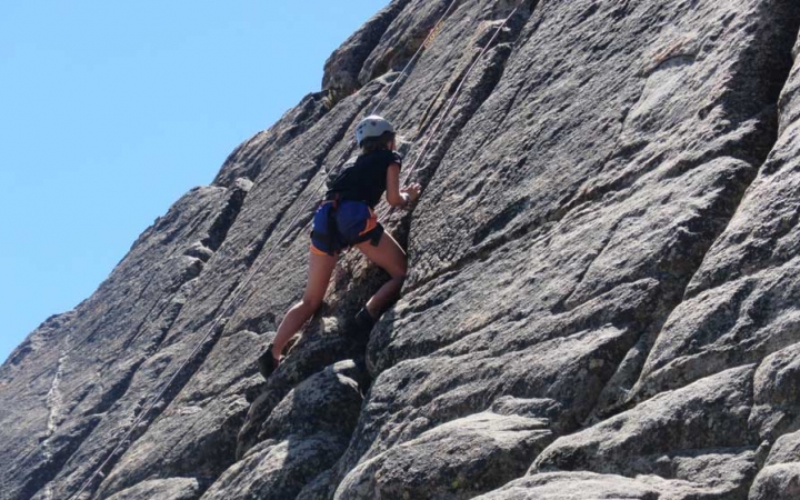 An outward bound student wearing safety gear climbs a rock wall under blue skies